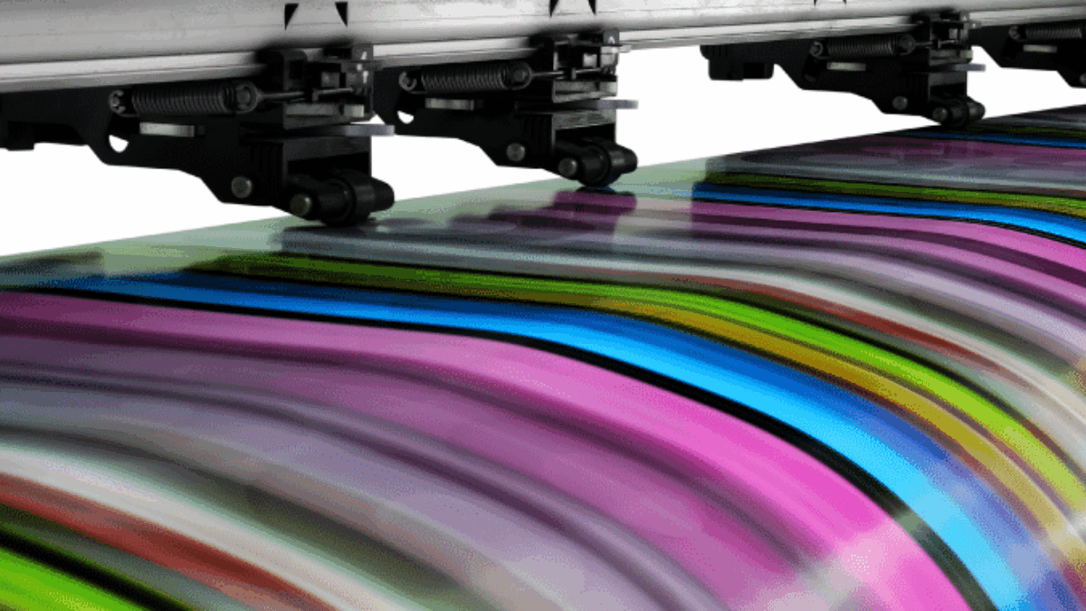 A close-up of a large-format printer producing vibrant, multicolored striped patterns on paper. The image shows the print head moving, with vivid hues of pink, blue, green, and yellow flowing across the paper.