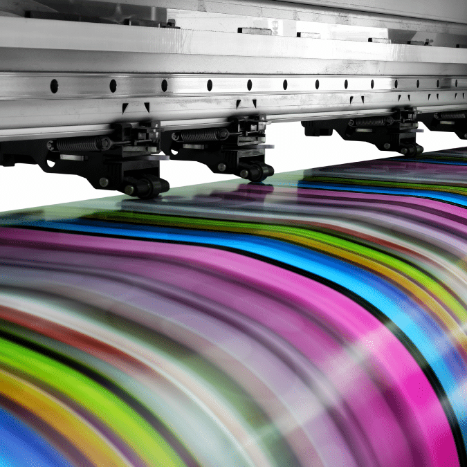 A close-up of a large-format printer producing vibrant, multicolored striped patterns on paper. The image shows the print head moving, with vivid hues of pink, blue, green, and yellow flowing across the paper.