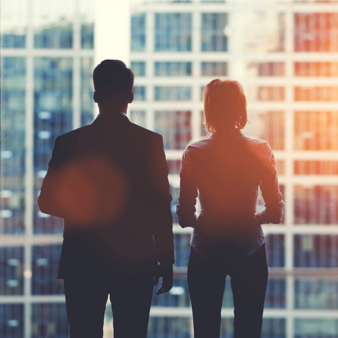 Two people in business attire stand side by side, silhouetted against a large office window with a cityscape of tall buildings in the background, sunlight streaming in.