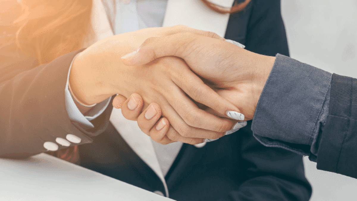 Two people in business attire shaking hands over a desk with documents, a pen, and eyeglasses, suggesting a successful meeting or agreement.