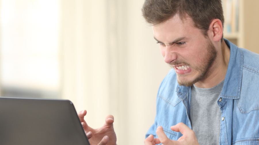 A young man sitting at a desk in front of a laptop clenches his fists and grimaces in frustration, appearing angry or stressed. Rolled-up papers and a coffee cup are on the desk.