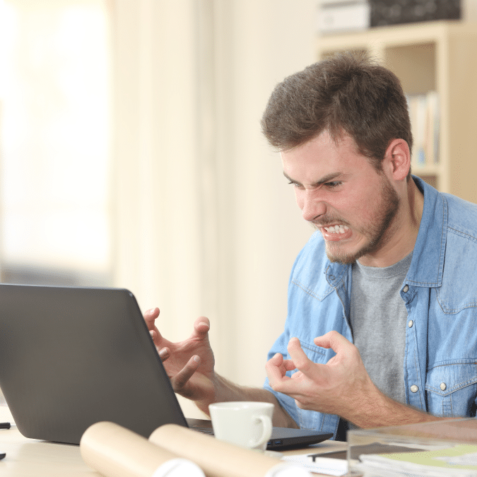 A young man sitting at a desk in front of a laptop clenches his fists and grimaces in frustration, appearing angry or stressed. Rolled-up papers and a coffee cup are on the desk.