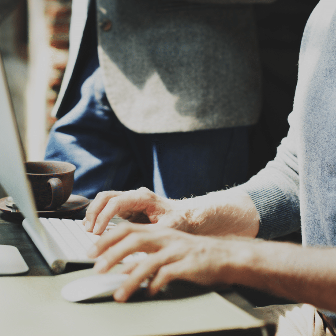 Two people working together at a desk; one is typing on a keyboard while the other stands nearby. A coffee cup is next to the computer. Only their hands and part of their bodies are visible.