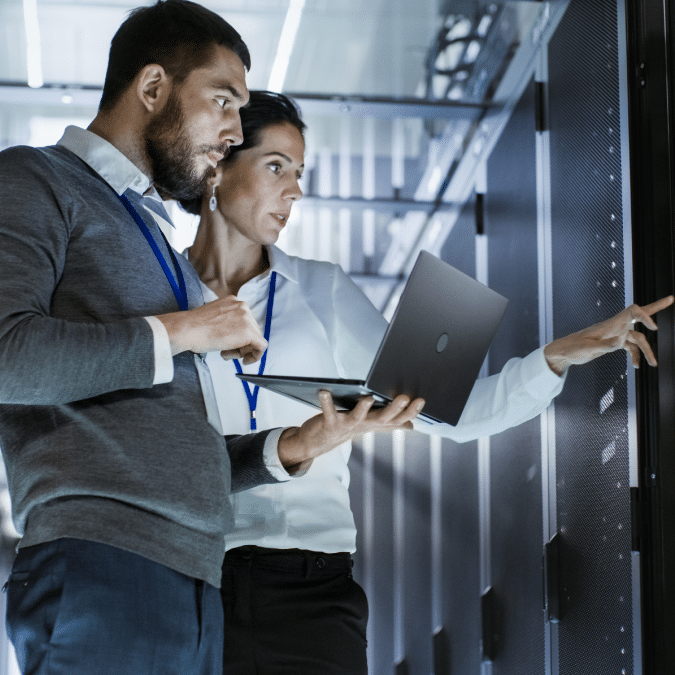 Two IT professionals in a server room, one holding a laptop and the other pointing at a server rack, both wearing ID badges and focused on their work.