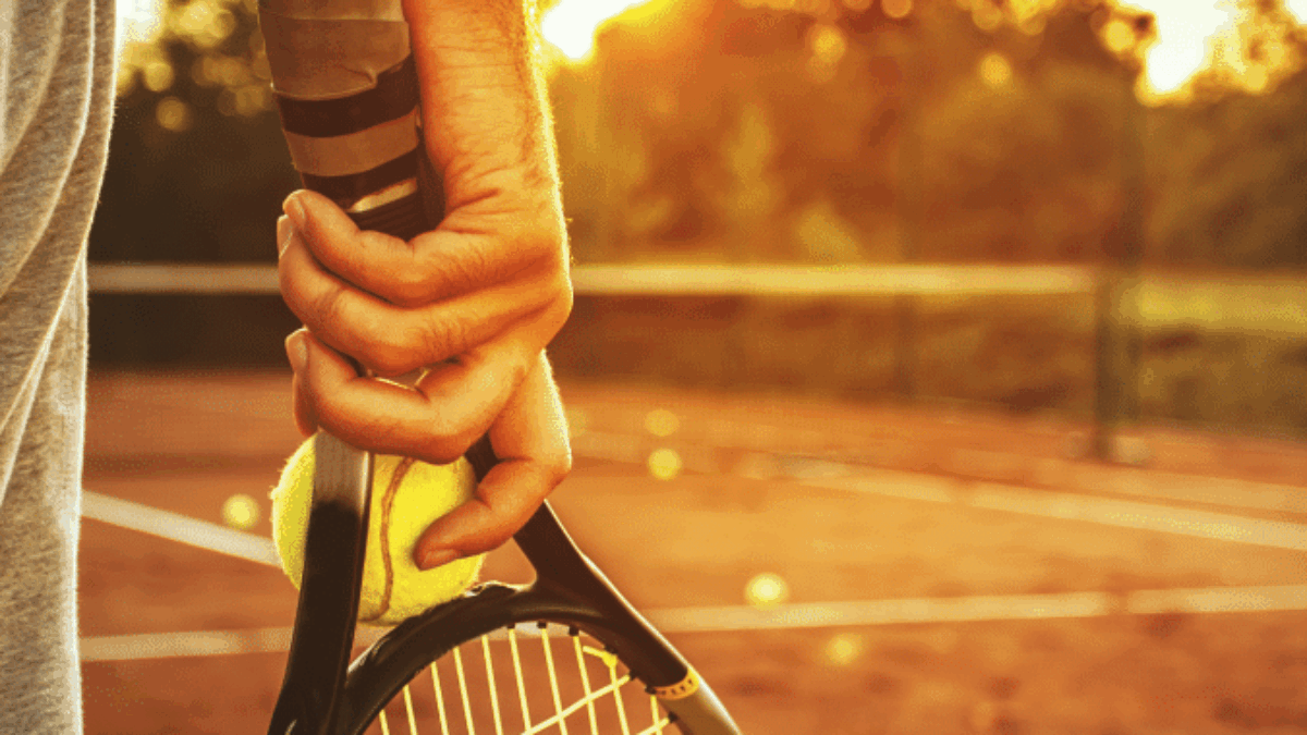 Close-up of a person’s hand holding a tennis racket and a yellow tennis ball on a clay tennis court at sunset, with a warm golden glow and blurred trees in the background.