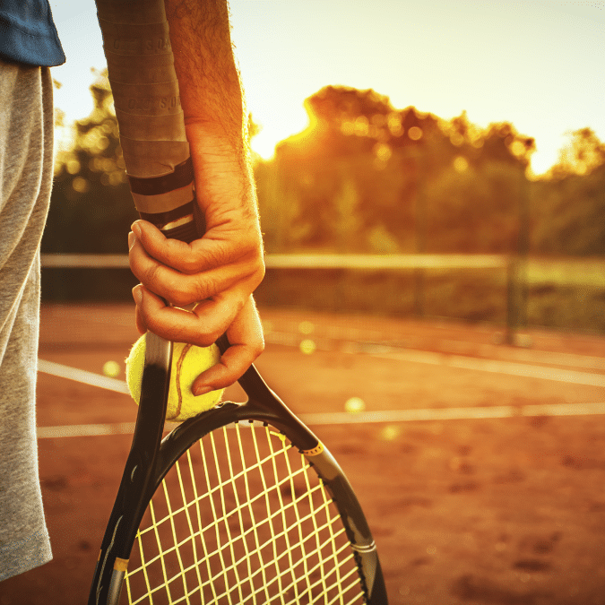 Close-up of a person’s hand holding a tennis racket and a yellow tennis ball on a clay tennis court at sunset, with a warm golden glow and blurred trees in the background.