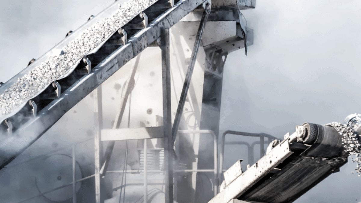 Industrial conveyor belts transport gravel amid clouds of dust at a quarry or mining site, with metal structures and machinery visible in the background.