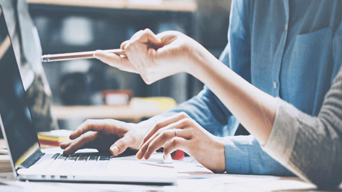 Two people work at a table with a laptop, papers, and pens. One person types on the keyboard while the other points at the screen with a pen, suggesting collaboration and discussion.