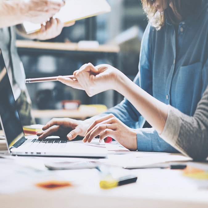 Two people work at a table with a laptop, papers, and pens. One person types on the keyboard while the other points at the screen with a pen, suggesting collaboration and discussion.