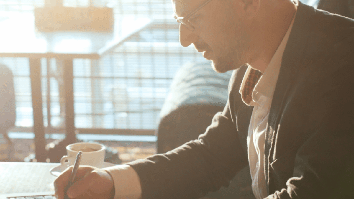 A man in a suit and glasses sits at a table in a sunlit cafe, writing in a notebook with a cup of coffee nearby and a laptop open in front of him.