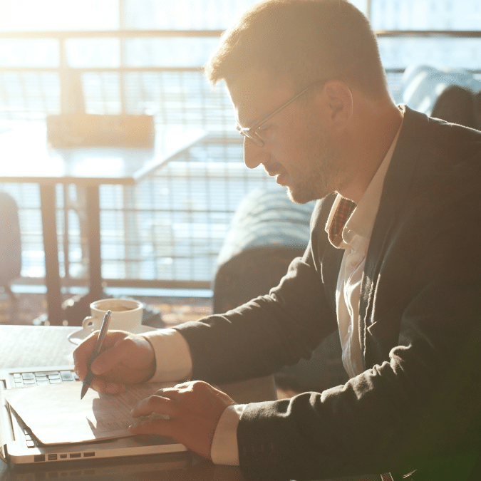 A man in a suit and glasses sits at a table in a sunlit cafe, writing in a notebook with a cup of coffee nearby and a laptop open in front of him.
