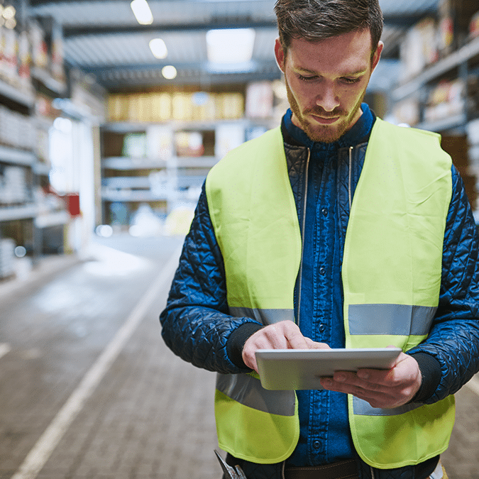 A man wearing a yellow safety vest stands in a warehouse, looking down and using a tablet. Shelves with goods are visible in the background, and the setting appears organized and well-lit.