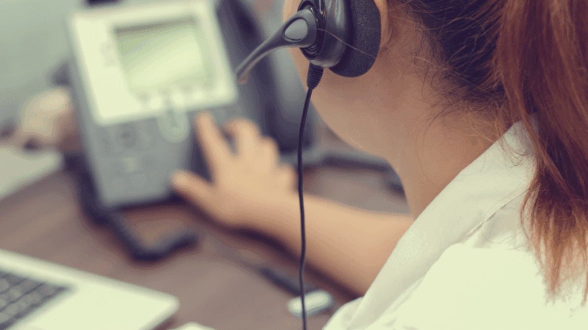 A woman wearing a headset sits at a desk, using a telephone while looking at a computer screen and holding a pen, suggesting she is working in a customer service or call center environment.