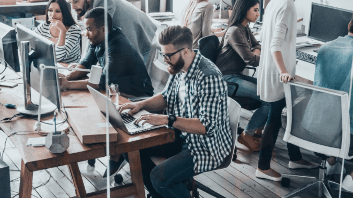Open-plan office with several young adults working at computers and collaborating in pairs, sitting at wooden desks with modern chairs and monitors, in a bright room with natural light.