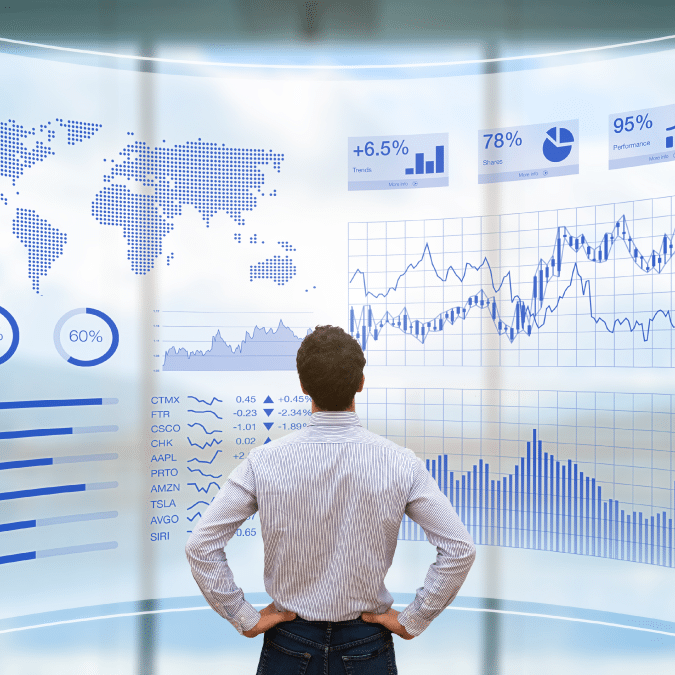 A man stands with hands on hips, facing a large digital screen displaying world maps, stock market data, charts, graphs, and various financial statistics in blue tones.