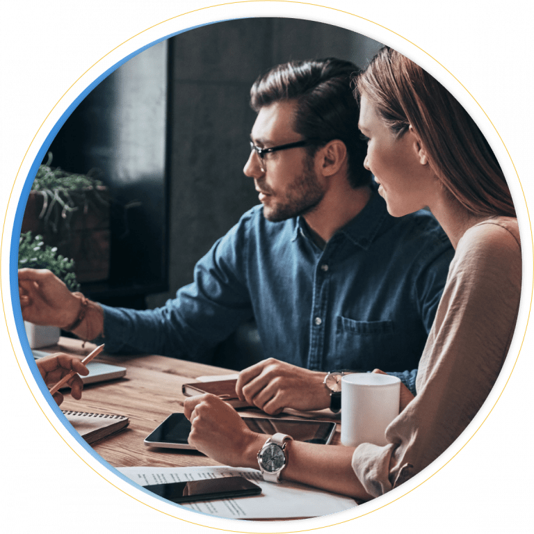 Two people sit at a wooden table with notebooks, a tablet, a smartphone, and a coffee mug, attentively discussing Dynamics 365 Marketing in a professional setting.