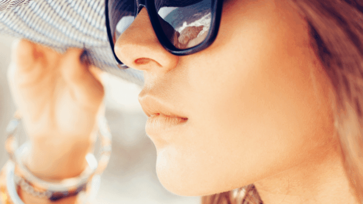 A woman wearing large black sunglasses and a wide-brimmed hat looks into the distance. She holds the brim of her hat with one hand, and colorful bracelets adorn her wrist. The image is bright and summery.