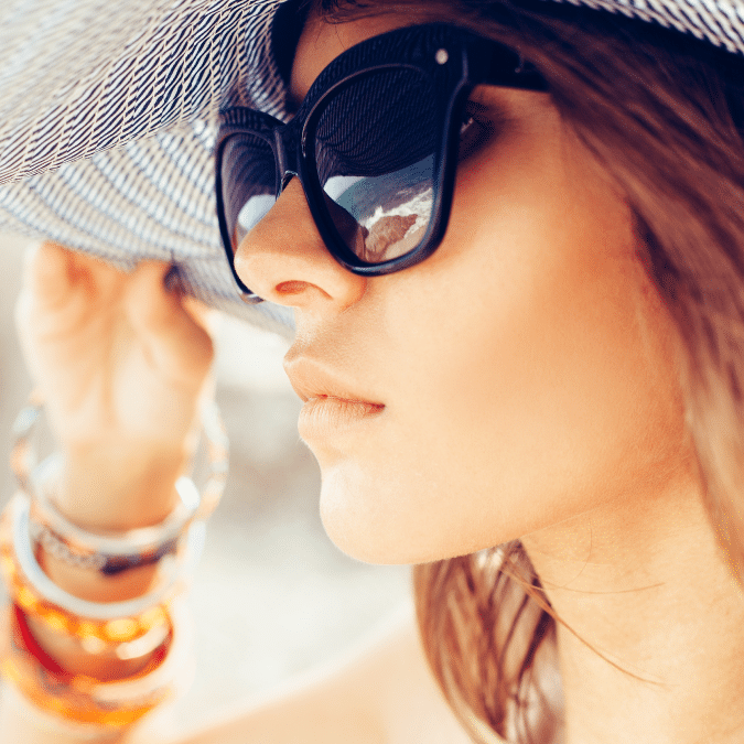 A woman wearing large black sunglasses and a wide-brimmed hat looks into the distance. She holds the brim of her hat with one hand, and colorful bracelets adorn her wrist. The image is bright and summery.