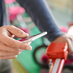 A person uses a smartphone to unlock or interact with a bike-sharing dock, with blurred bikes and bright colors in the background.