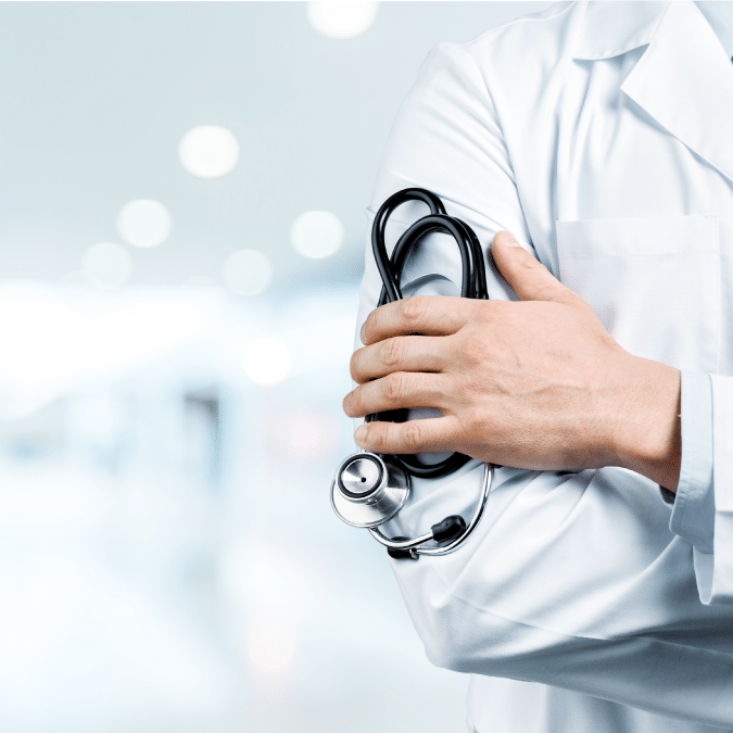 A doctor wearing a white lab coat stands with arms crossed, holding a stethoscope in one hand. The background is blurred, suggesting a hospital or medical facility setting.