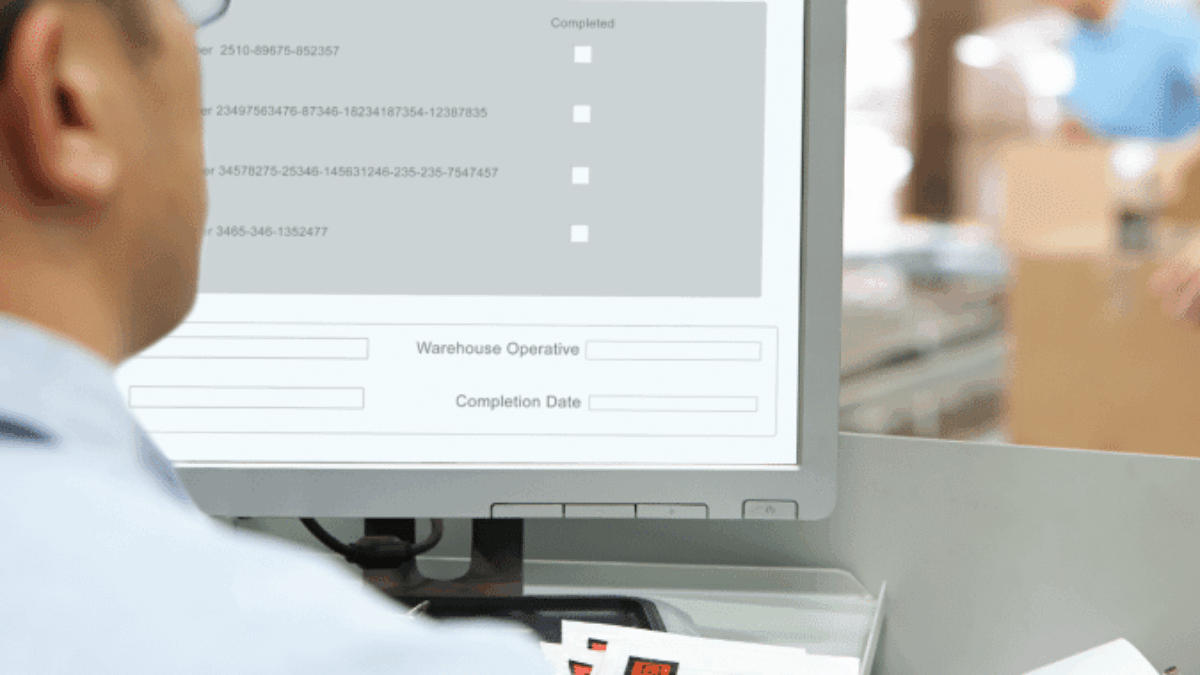 A person wearing glasses is sitting at a desk and looking at a computer screen displaying a warehouse management system. Stacks of papers and boxes are visible nearby, with another person blurred in the background.