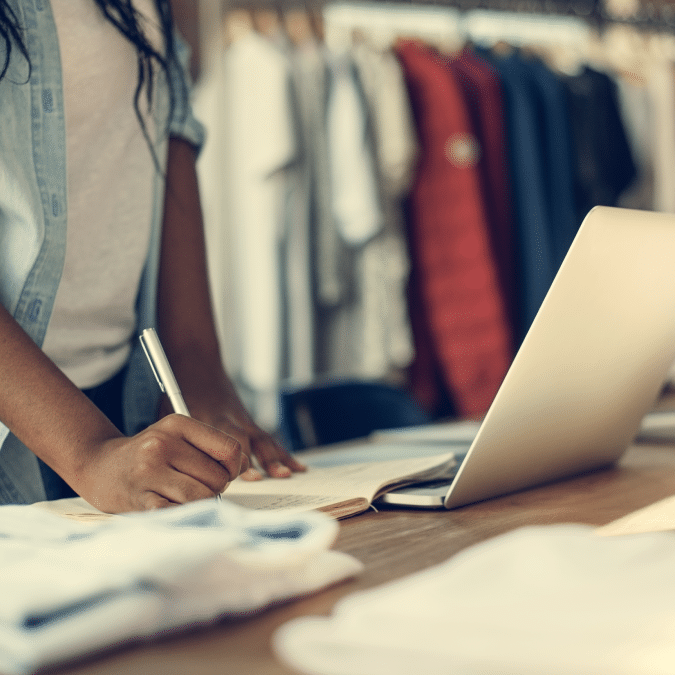 A person writes in a notebook with one hand while standing at a table with a laptop open, surrounded by neatly folded clothes in a retail store. Clothing hangs in the background.