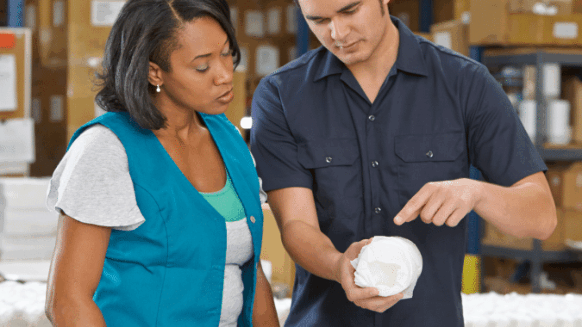 Two workers in a warehouse examine a rolled-up product. The man points at the item while the woman looks on attentively. Shelves with boxes and other items are visible in the background.