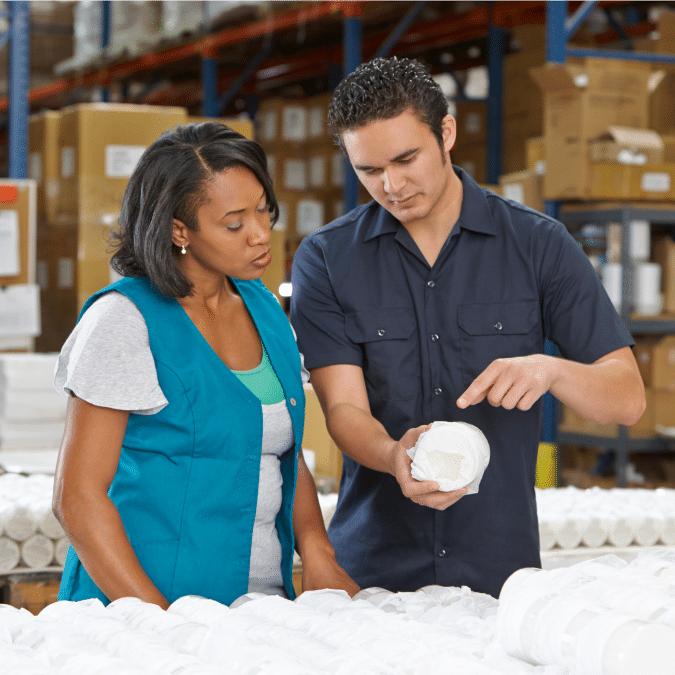 Two workers in a warehouse examine a rolled-up product. The man points at the item while the woman looks on attentively. Shelves with boxes and other items are visible in the background.