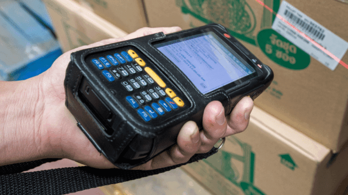 A person scans a barcode on a cardboard box using a handheld barcode scanner in a warehouse setting.
