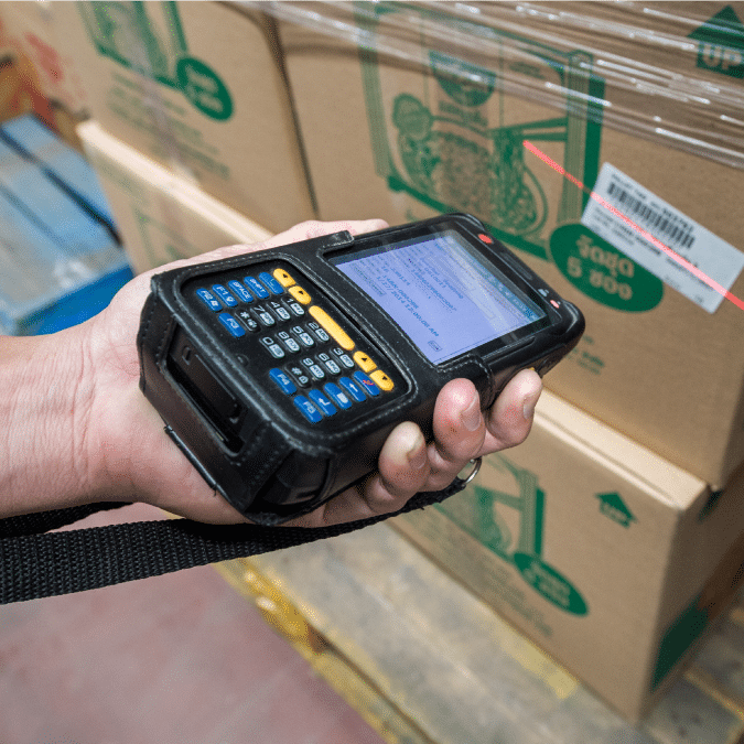 A person scans a barcode on a cardboard box using a handheld barcode scanner in a warehouse setting.