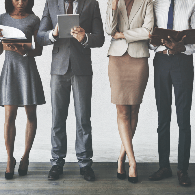 Four professionally dressed people stand side by side, holding journals and digital devices, with only their lower faces and bodies visible, suggesting a business or office setting.