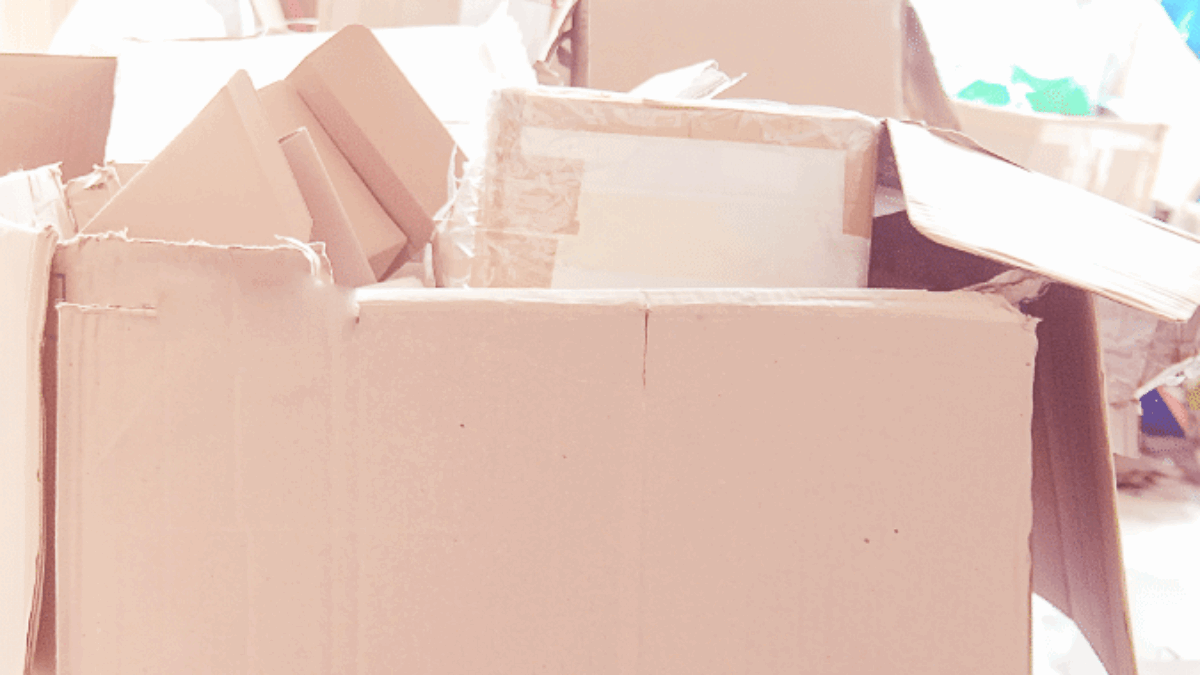 A large cardboard box, partially open and filled with packing materials and smaller boxes, sits on a light-colored floor. Additional boxes and bags are scattered in the blurry background.