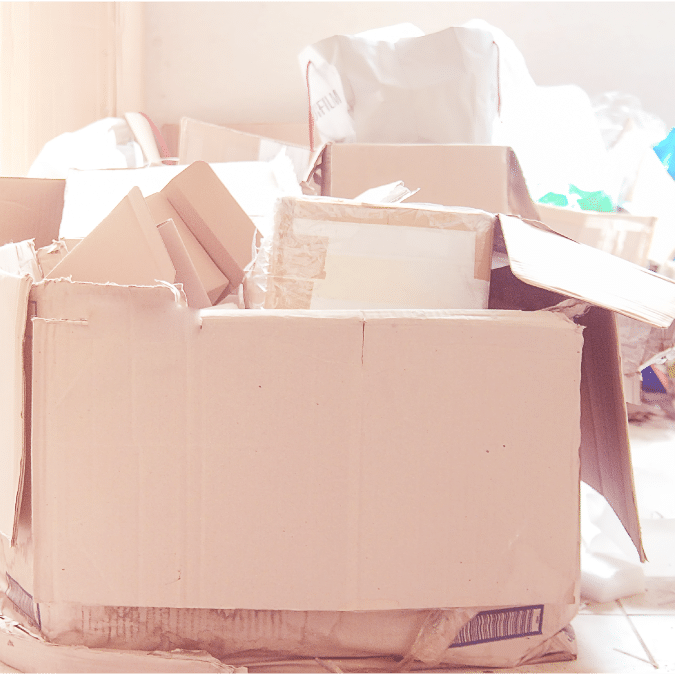 A large cardboard box, partially open and filled with packing materials and smaller boxes, sits on a light-colored floor. Additional boxes and bags are scattered in the blurry background.