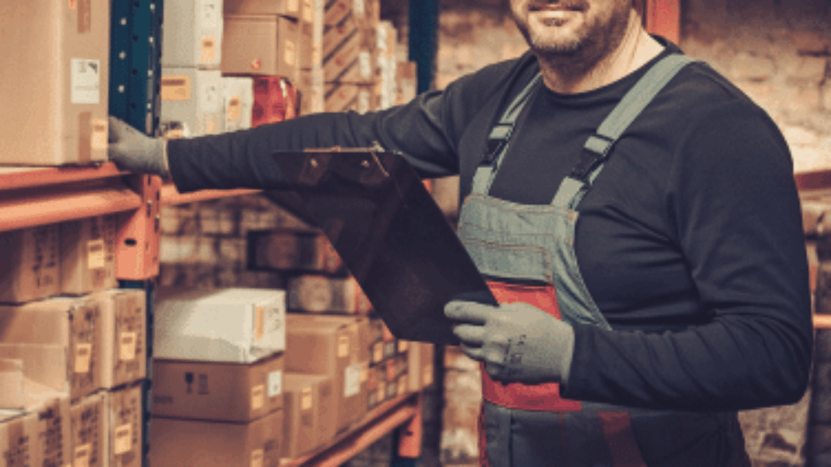 A warehouse worker wearing gloves and overalls holds a clipboard while standing next to shelves filled with cardboard boxes. He is smiling and appears to be checking inventory.