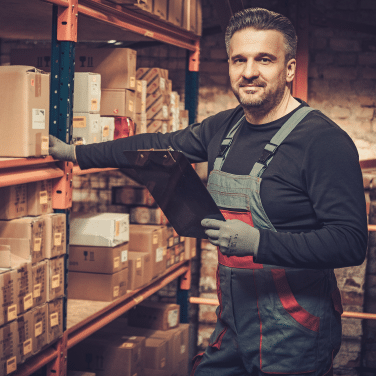 A warehouse worker wearing gloves and overalls holds a clipboard while standing next to shelves filled with cardboard boxes. He is smiling and appears to be checking inventory.