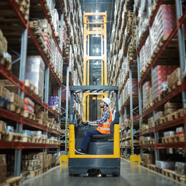 A worker in a safety vest and helmet operates a forklift between tall shelves stacked with boxes in a large warehouse.