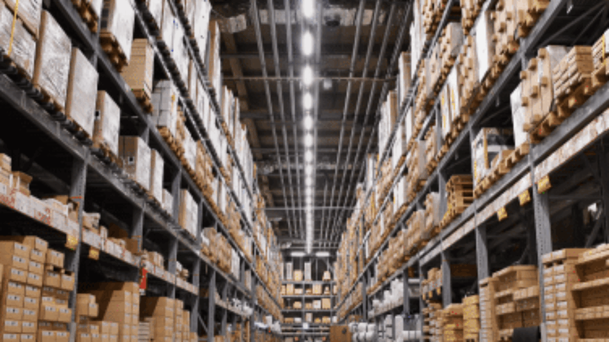 A wide-angle view of a large warehouse with tall shelves stacked with various boxes and packages, under bright overhead lighting, and a shiny polished floor.