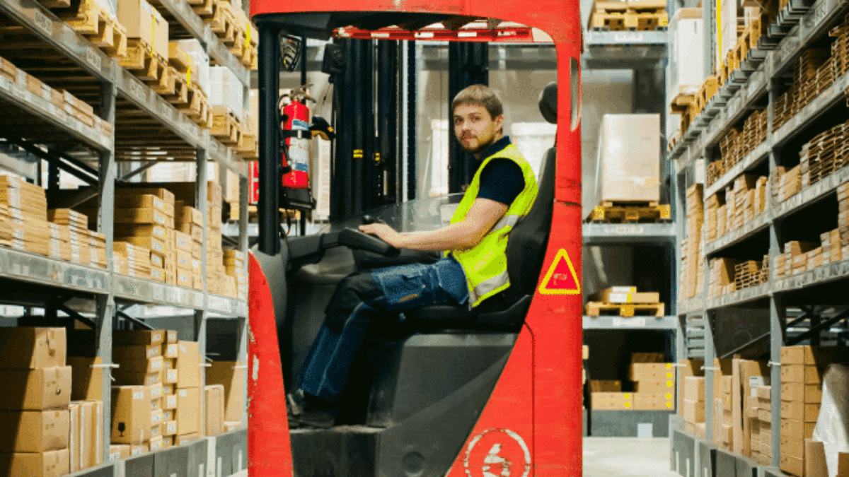 A man wearing a yellow safety vest operates a red forklift in a warehouse aisle filled with neatly stacked cardboard boxes on metal shelves.