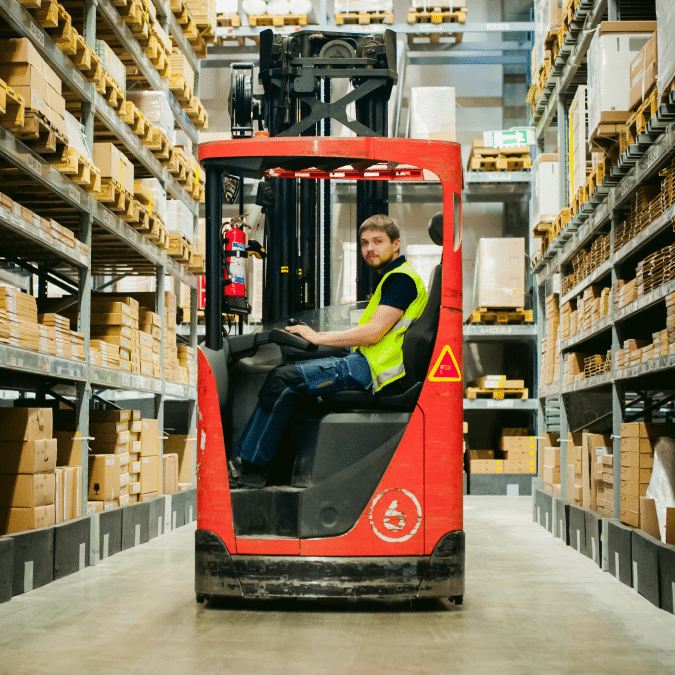 A man wearing a yellow safety vest operates a red forklift in a warehouse aisle filled with neatly stacked cardboard boxes on metal shelves.