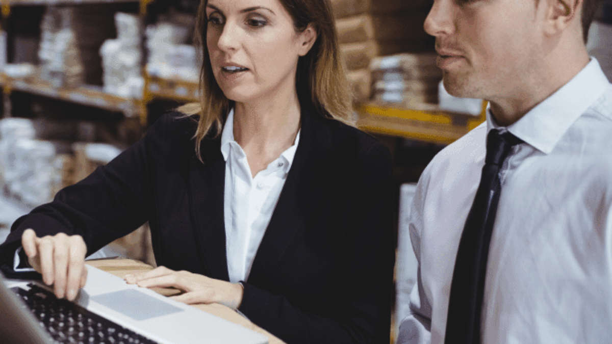 Two people in business attire look at a laptop in a warehouse, surrounded by shelves stacked with boxes and packages. One person is typing while the other observes attentively.