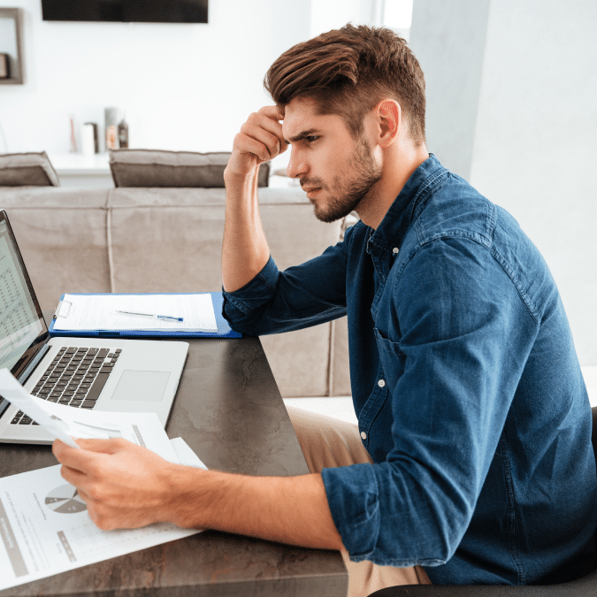 A man sits at a table looking stressed while reviewing documents and charts in front of a laptop, with a clipboard and pen on the table in a bright, modern living room.