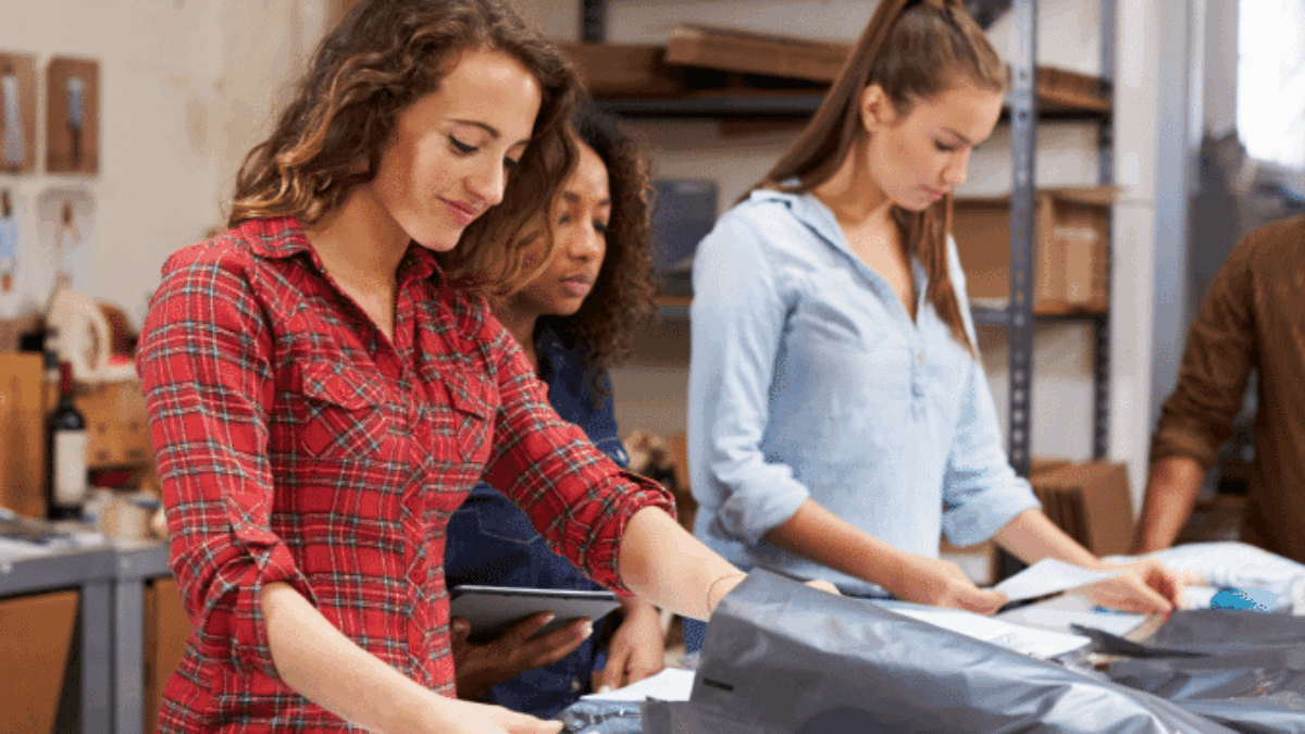 Three women stand at a table in a workshop or warehouse, sorting and folding clothes. Shelves with boxes and supplies are in the background. They appear focused on their task.