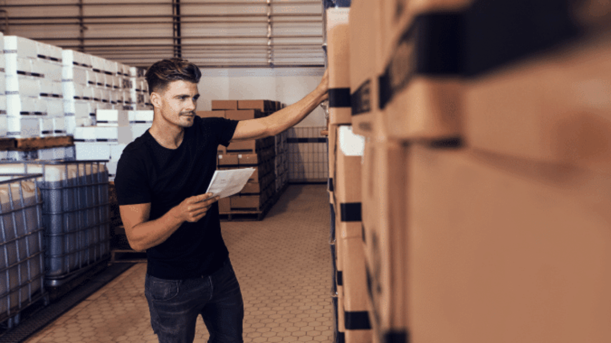 A man in a warehouse holds a clipboard and inspects stacked boxes, appearing to check inventory. He wears a black t-shirt and jeans, and the warehouse has hexagonal floor tiles and metal shelving.