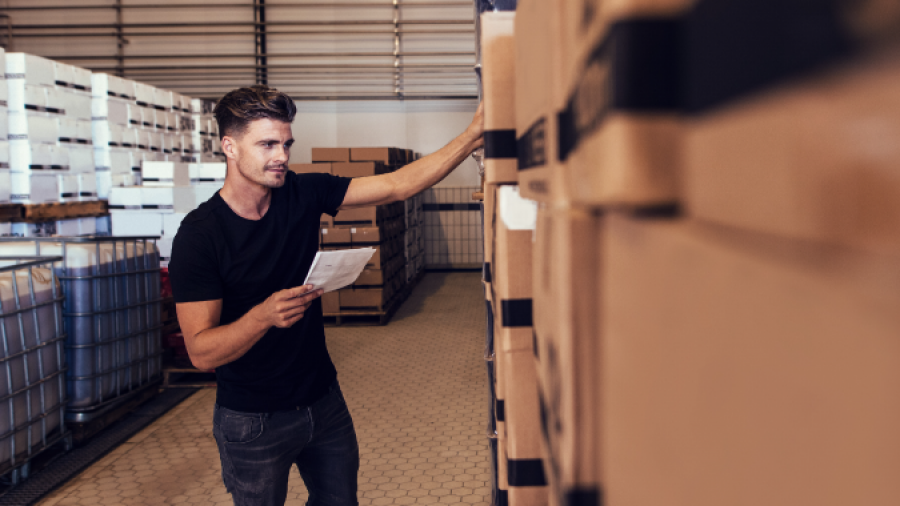 A man in a warehouse holds a clipboard and inspects stacked boxes, appearing to check inventory. He wears a black t-shirt and jeans, and the warehouse has hexagonal floor tiles and metal shelving.