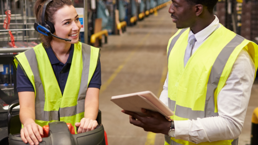 A woman wearing a headset and high-visibility vest sits on a forklift, smiling and talking to a man in a safety vest holding a tablet in a large warehouse.