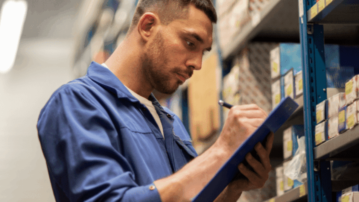 A man in a blue work uniform writes on a clipboard while standing beside shelves filled with boxes and parts in a warehouse or storage facility.