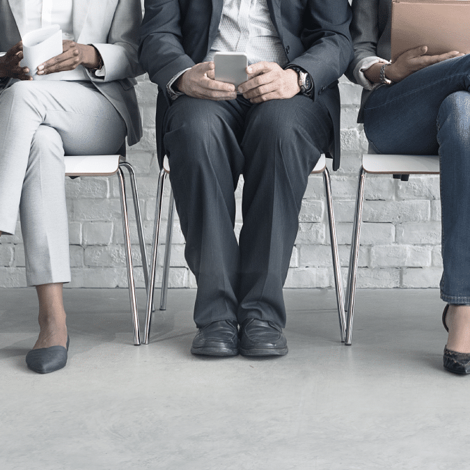 Three people in business attire sit on chairs against a white brick wall, each holding a smartphone or tablet, with only their lower bodies visible.
