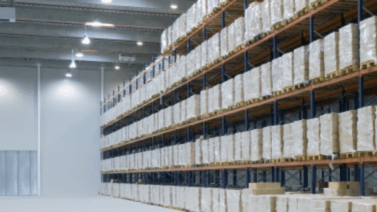 A spacious warehouse with tall shelves stacked neatly with pallets of boxes, illuminated by overhead lights. The floor is clean and mostly empty.