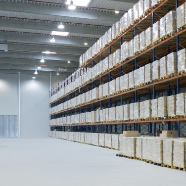 A spacious warehouse with tall shelves stacked neatly with pallets of boxes, illuminated by overhead lights. The floor is clean and mostly empty.
