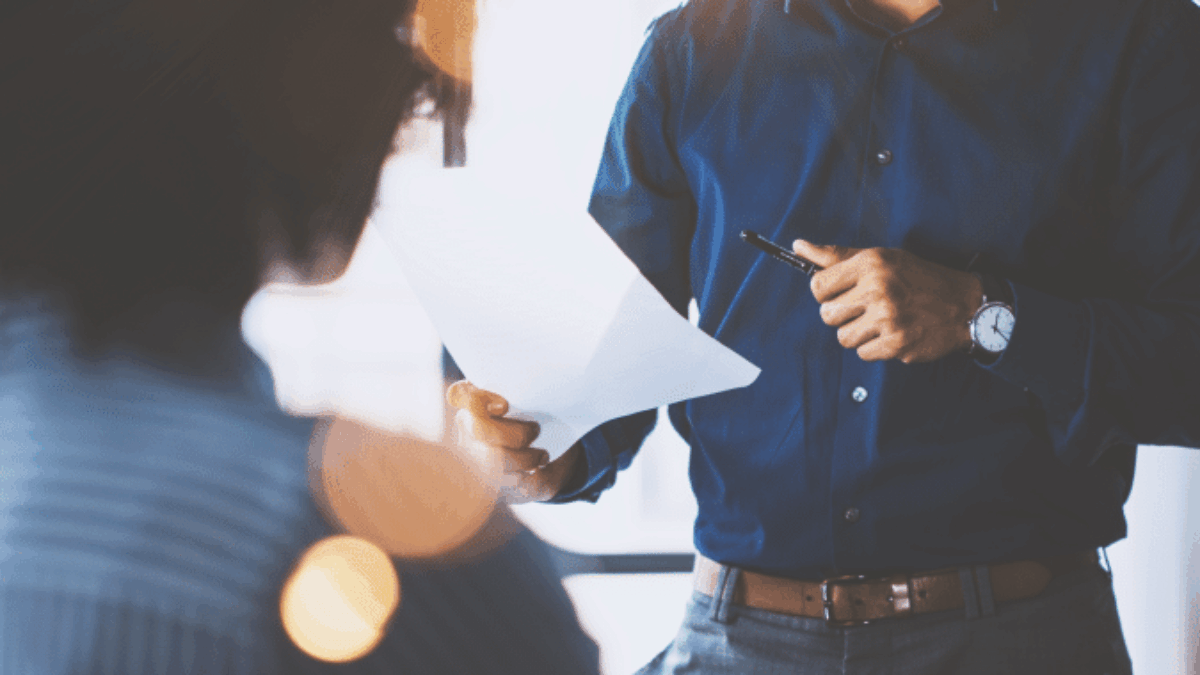 A man in a blue shirt stands holding papers and a pen, smiling as he talks to a seated person in an office setting with sunlight streaming in.